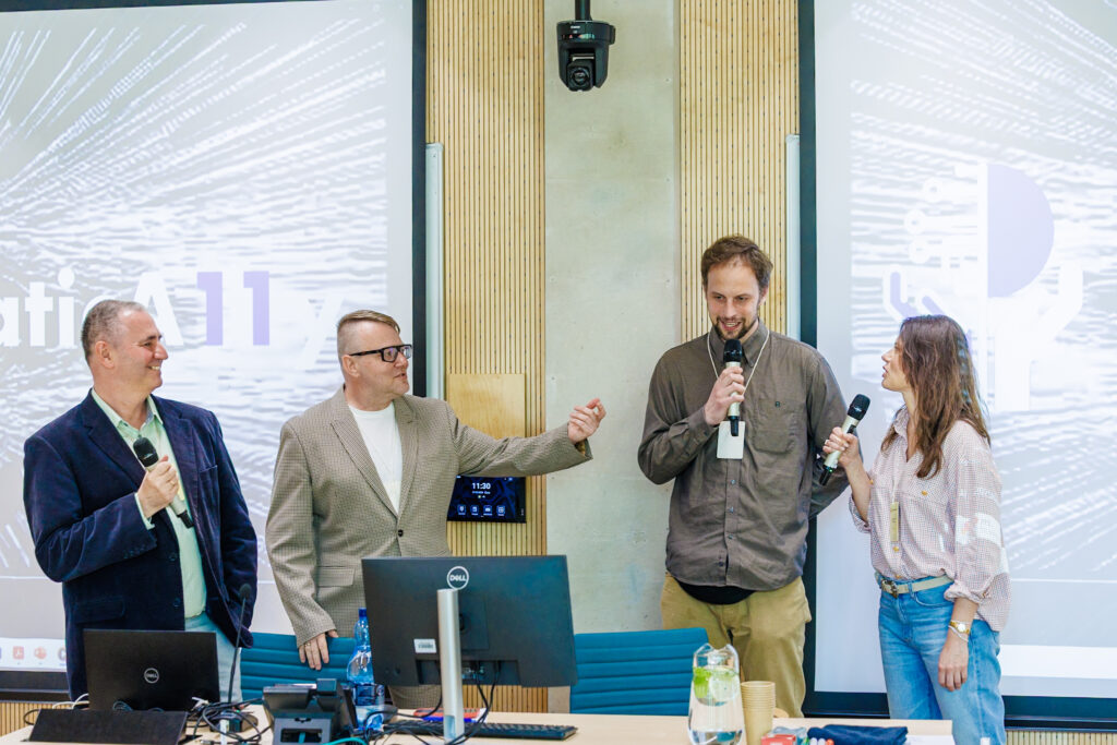 Czworo prowadzących (dwóch mężczyzn, dwie kobiety) stoi razem na scenie, część trzyma mikrofony, w tle dwa ekrany – moment panelu dyskusyjnego.