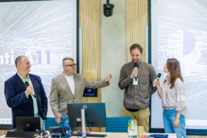 Czworo prowadzących (dwóch mężczyzn, dwie kobiety) stoi razem na scenie, część trzyma mikrofony, w tle dwa ekrany – moment panelu dyskusyjnego.