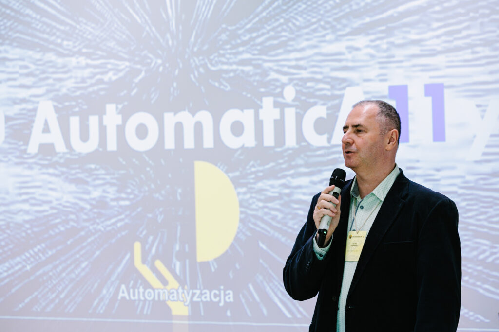 Jacek Zadrożny w ciemnej marynarce przemawia do mikrofonu przed dużym ekranem z logo AutomaticAlly, widoczny identyfikator z imieniem i nazwiskiem.