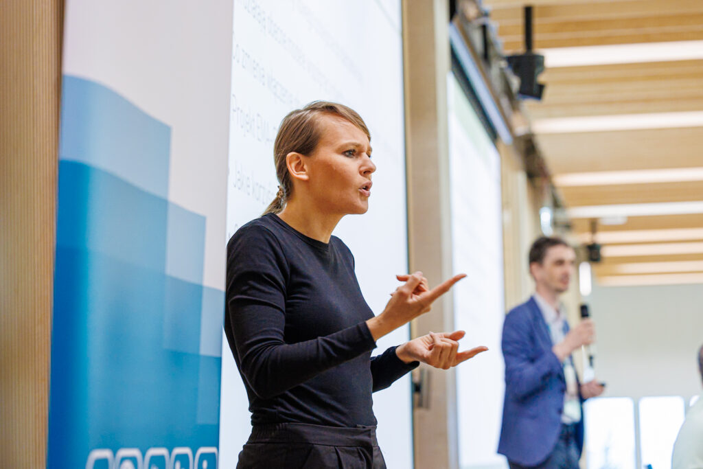 Tłumaczka języka migowego w czarnym stroju pokazuje znak migowy przed ekranem z listą punktów prezentacji.