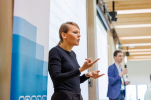 Tłumaczka języka migowego w czarnym stroju pokazuje znak migowy przed ekranem z listą punktów prezentacji.