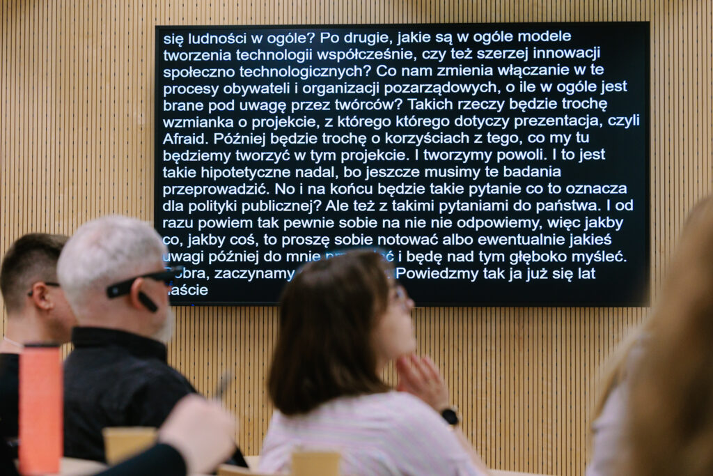 Ekran z automatyczną transkrypcją na żywo wyświetlaną podczas prezentacji, widoczni słuchacze na widowni.