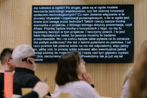 Ekran z automatyczną transkrypcją na żywo wyświetlaną podczas prezentacji, widoczni słuchacze na widowni.