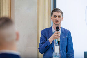 Zbliżenie twarzy mężczyzny w niebieskiej marynarce przemawiającego do mikrofonu, z identyfikatorem konferencyjnym na smyczy, w tle jasny ekran prezentacji.