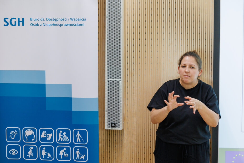 Tłumaczka języka migowego w czarnym stroju pokazuje znak migowy, w tle baner SGH Biuro ds. Dostępności i Wsparcia Osób z Niepełnosprawnościami z piktogramami dostępności.