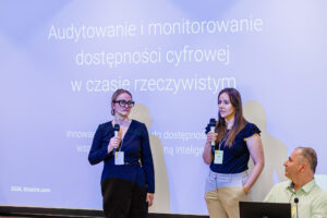 Dwie prowadzące w granatowych strojach przemawiają do mikrofonów przed ekranem z tytułem "Audytowanie i monitorowanie dostępności cyfrowej w czasie rzeczywistym", obok siedzi mężczyzna ze słuchawką douszną.