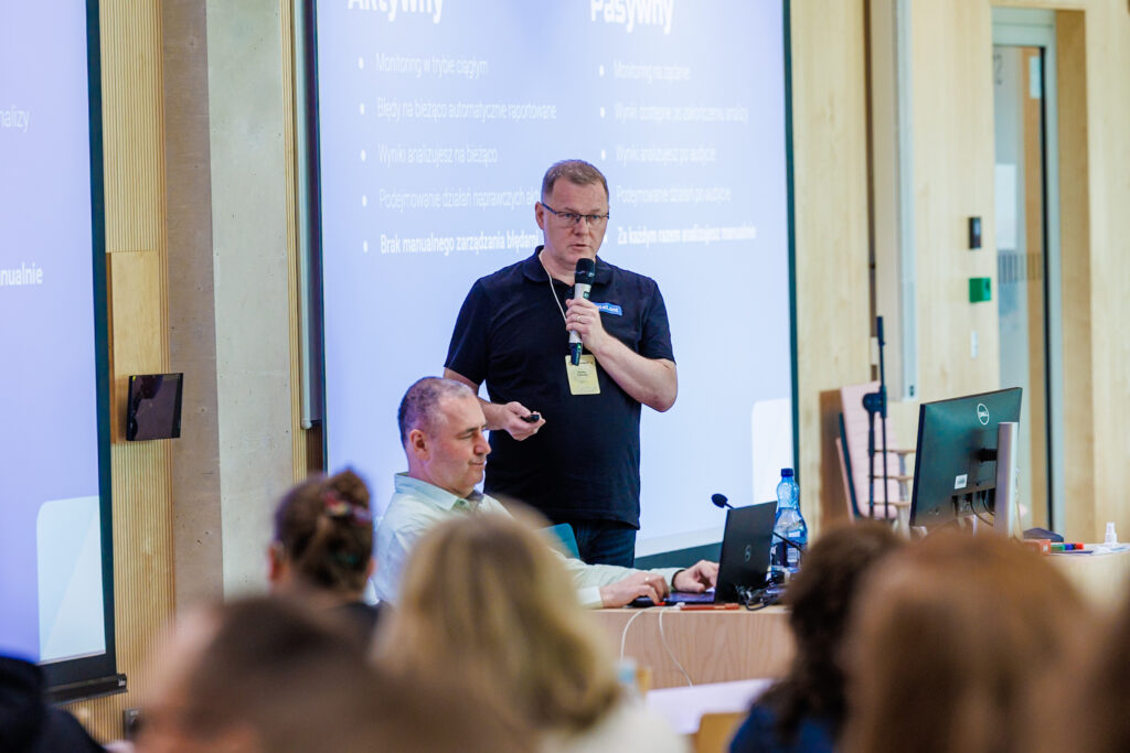 Cezary Tomczyk przemawia do mikrofonu przed slajdem porównującym monitoring aktywny i pasywny, obok mężczyzna pracujący na laptopie.