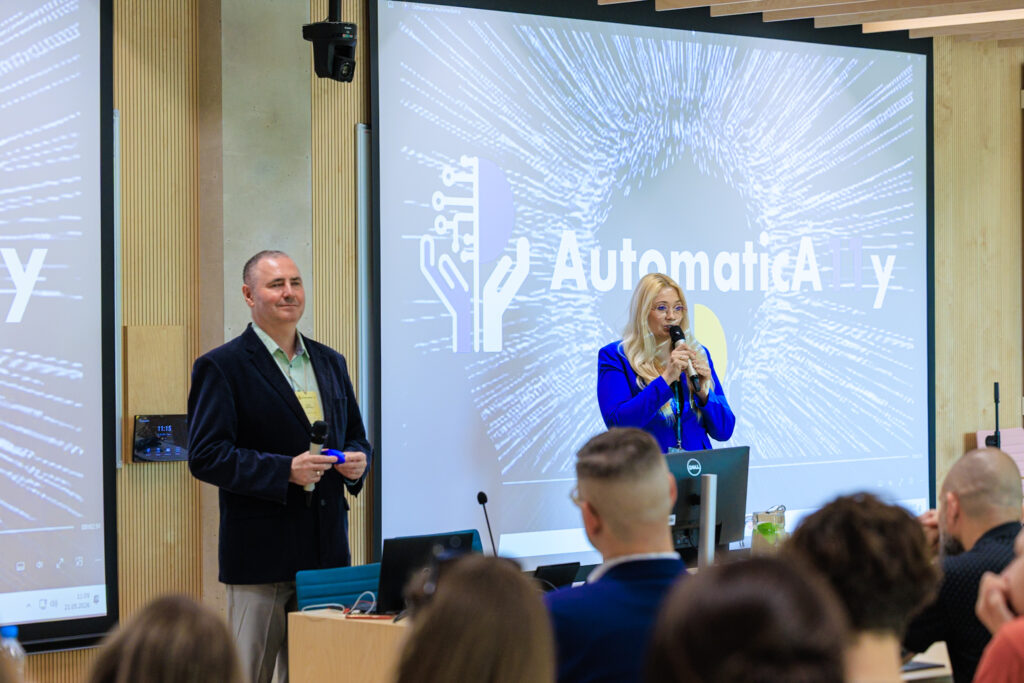 Otwarcie konferencji – prowadzący w granatowej marynarce i prowadząca w niebieskiej marynarce z mikrofonem stoją przed ekranem z logo AutomaticAlly.
