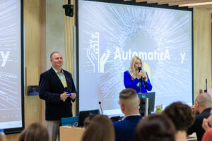 Otwarcie konferencji – prowadzący w granatowej marynarce i prowadząca w niebieskiej marynarce z mikrofonem stoją przed ekranem z logo AutomaticAlly.