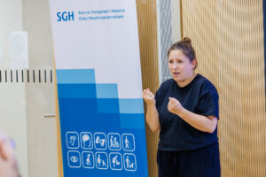 Tłumaczka języka migowego w czarnym stroju pokazuje znak migowy z zaciśniętymi pięściami, w tle baner SGH Biuro ds. Dostępności i Wsparcia Osób z Niepełnosprawnościami.
