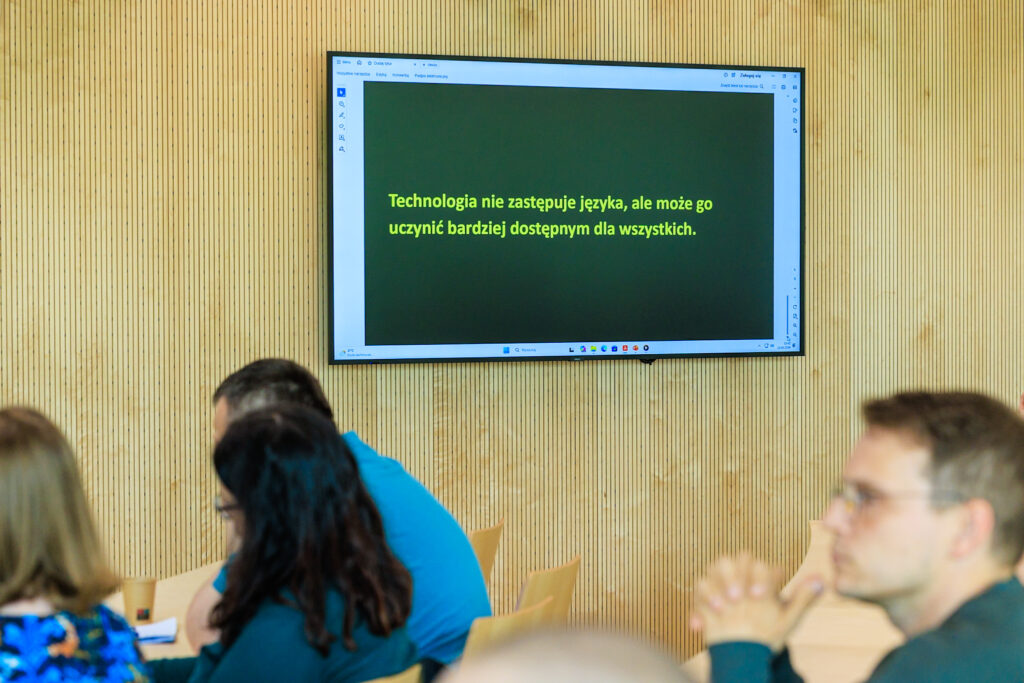 Ekran telewizyjny wyświetlający ten sam cytat o technologii i języku, na pierwszym planie rozmyci uczestnicy siedzący przy stole.