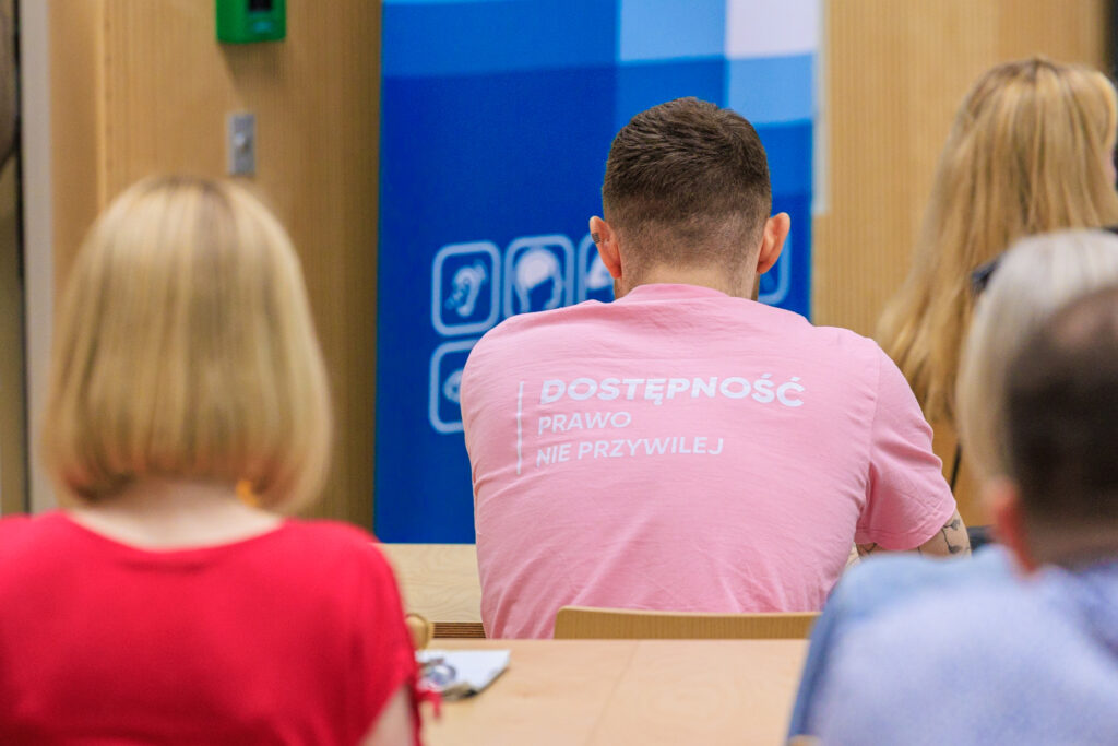 Mężczyzna widziany od tyłu w różowej koszulce z napisem "Dostępność, prawo, nie przywilej", w tle baner z piktogramami dostępności.
