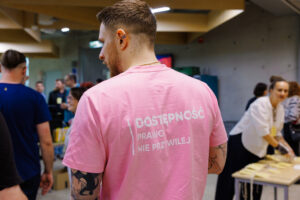 Mężczyzna widziany od tyłu w różowej koszulce z napisem "Dostępność, prawo, nie przywilej" i tatuażami na ramieniu, w tle inni uczestnicy przy stoiskach.