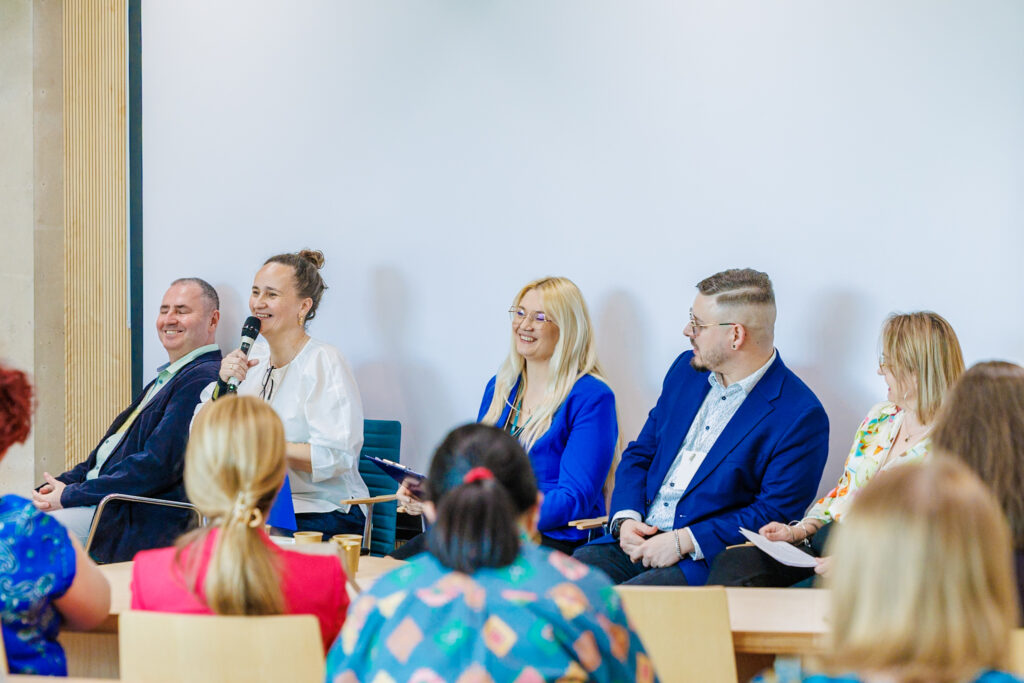 Panel dyskusyjny – pięcioro prelegentów siedzi w rzędzie, kobieta w białej bluzce mówi do mikrofonu, pozostali z uśmiechem słuchają.