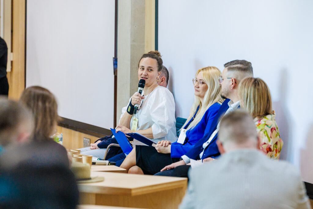 Panel dyskusyjny z bliska – kobieta w białej bluzce przemawia do mikrofonu, obok blondwłosa kobieta w niebieskiej marynarce i mężczyzna w niebieskim garniturze.