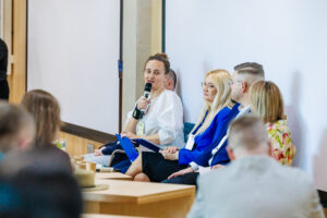 Panel dyskusyjny z bliska – kobieta w białej bluzce przemawia do mikrofonu, obok blondwłosa kobieta w niebieskiej marynarce i mężczyzna w niebieskim garniturze.