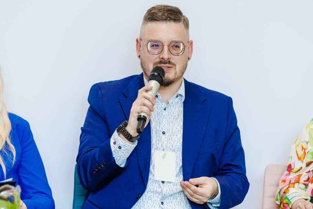 Mężczyzna w niebieskim garniturze i okularach przemawia do mikrofonu podczas panelu dyskusyjnego, z identyfikatorem konferencyjnym na smyczy.