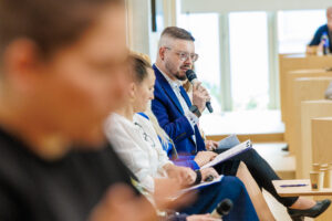 Mężczyzna w niebieskim garniturze przemawia do mikrofonu obok blondwłosej uczestniczki panelu trzymającej notatki.