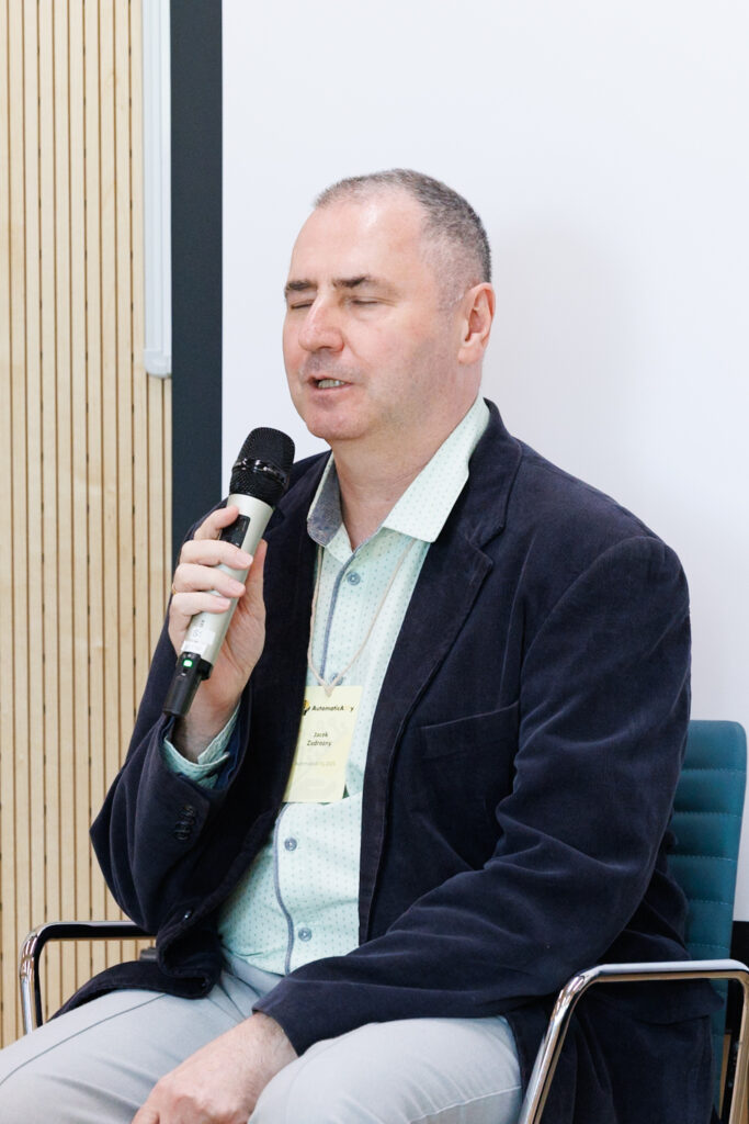 Jacek Zadrożny z zamkniętymi oczami przemawia do mikrofonu, w granatowej marynarce sztruksowej, z identyfikatorem konferencyjnym AutomaticAlly.