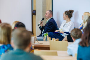 Jacek Zadrożny przemawia do mikrofonu podczas panelu dyskusyjnego, obok kobieta w białej bluzie z identyfikatorem AutomaticAlly i notatnikiem.