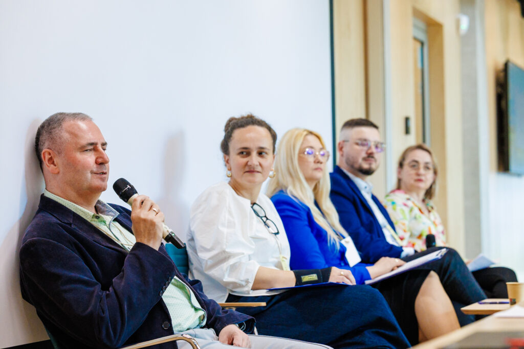 Panel dyskusyjny – mężczyzna w granatowej marynarce przemawia do mikrofonu, obok czworo prelegentów: kobieta w białej bluzce, blondwłosa kobieta w niebieskiej marynarce, mężczyzna w niebieskim garniturze i kobieta w kwiecistej bluzce.