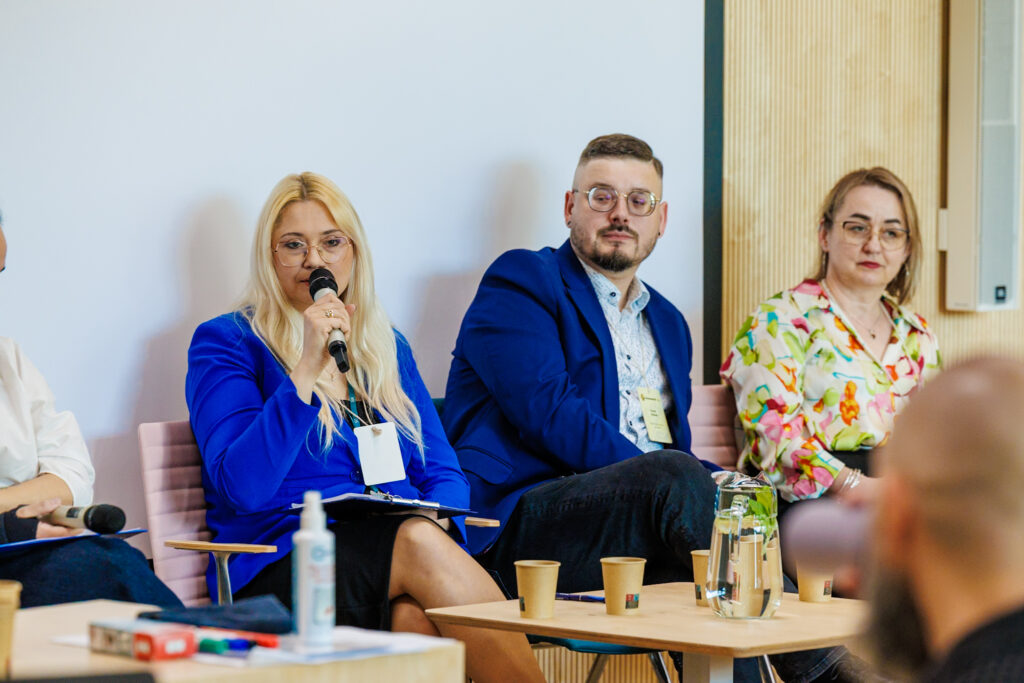 Blondwłosa kobieta w niebieskiej marynarce przemawia do mikrofonu podczas panelu, obok mężczyzna w okrągłych okularach i granatowej marynarce oraz kobieta w kwiecistej bluzce.