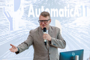 Mężczyzna w marynarce w kratę i okularach gestykuluje, przemawiając przed ekranem z logo konferencji.